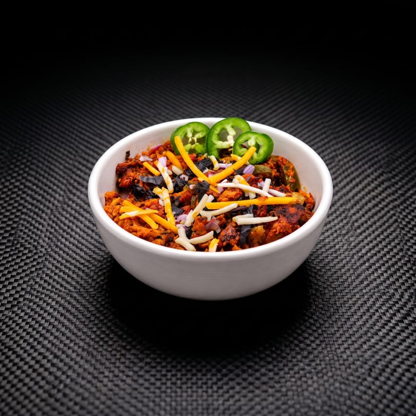 A white bowl filled with chili, topped with shredded cheese, diced onions, and jalapeño slices.