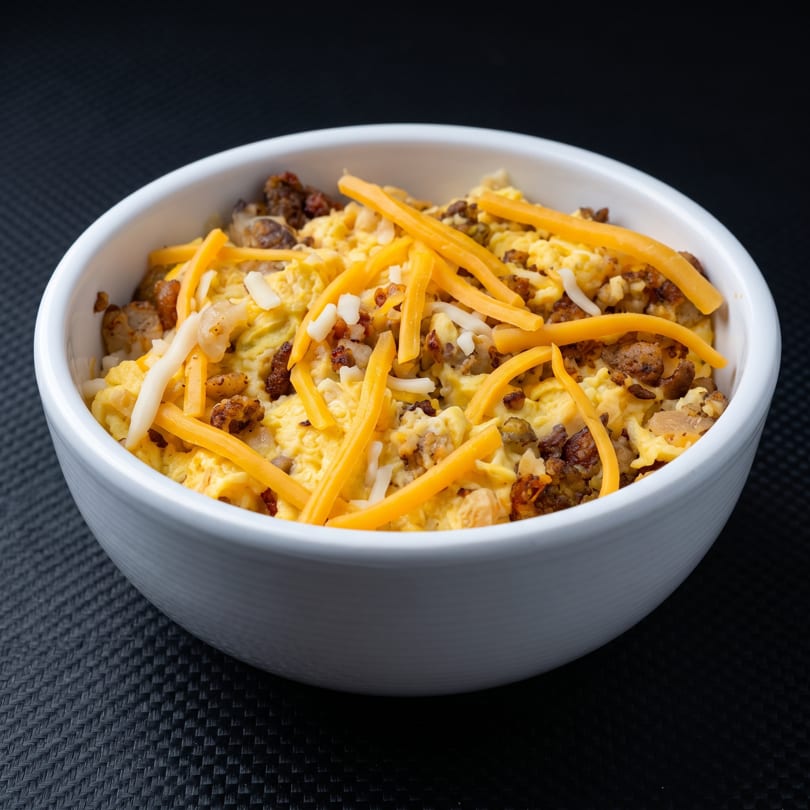 A white bowl filled with scrambled eggs, sausage crumbles, and shredded cheddar and white cheese.